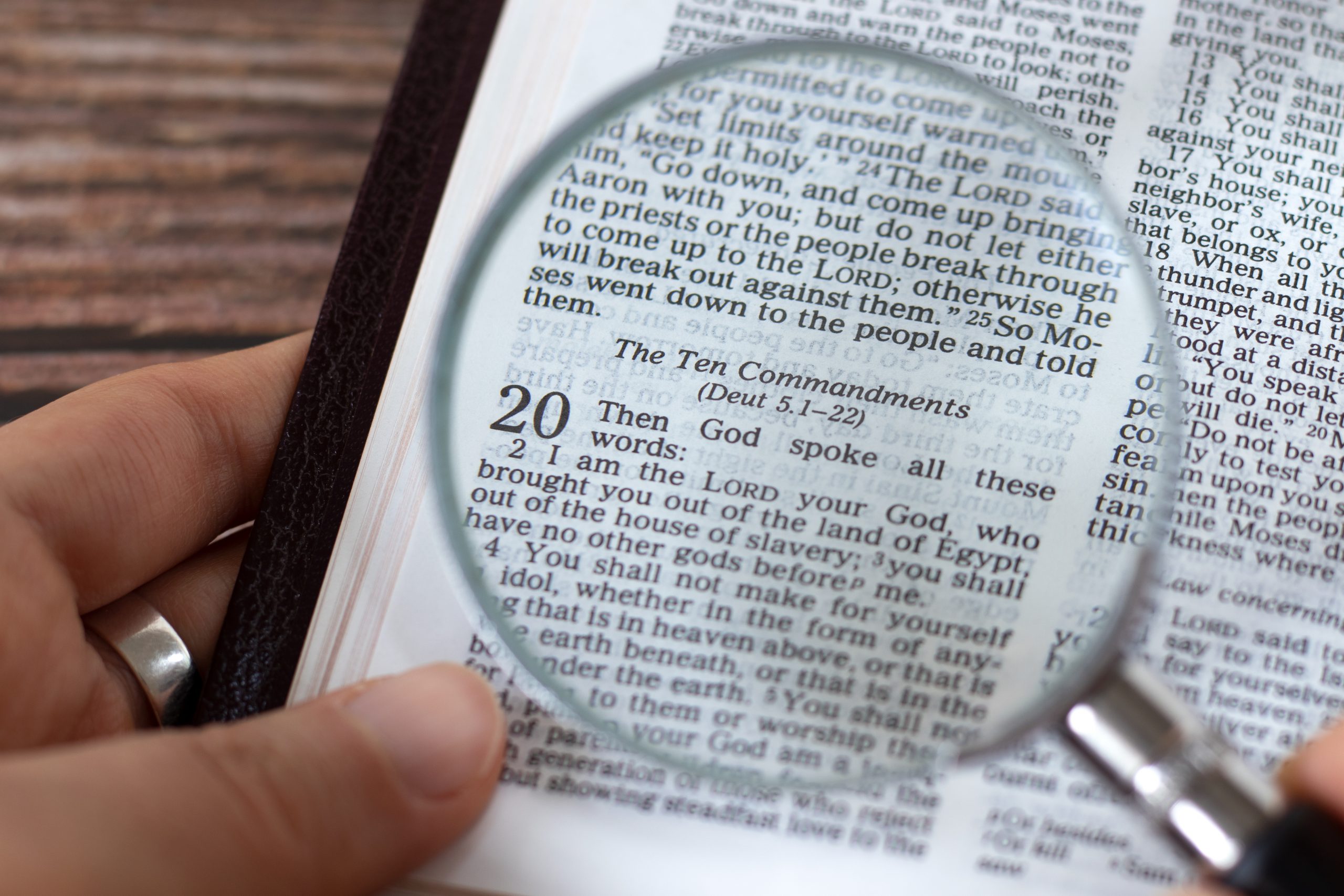 Human hand holding a magnifying glass over open holy bible book of Exodus verses for Ten Commandments, top view